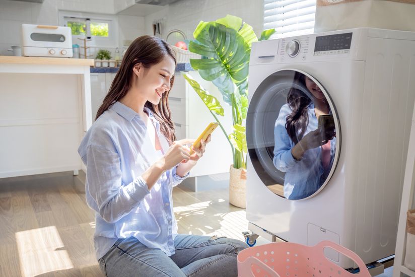 Woman looks at her smartphone while seated in front of her washing machine.